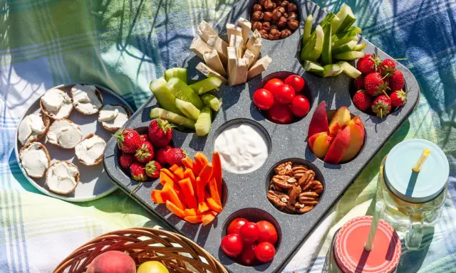 Leckere Picknick-Snacks: Erdbeeren, Gurken, Karotten, Nüsse, Tomaten, Pfirsiche und Brotscheiben mit Frischkäse auf einer Decke.