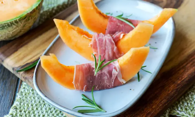 Melone mit Schinken, garniert mit Rosmarin, auf einem Teller angerichtet. Ein halber Melone liegt daneben auf einem Holzbrett.