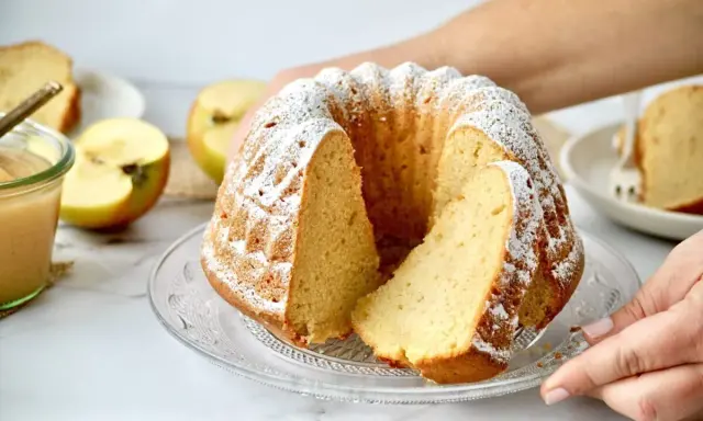 Saftiger Apfelkuchen mit Puderzucker, daneben ein Glas Apfelmus und frische Äpfel.