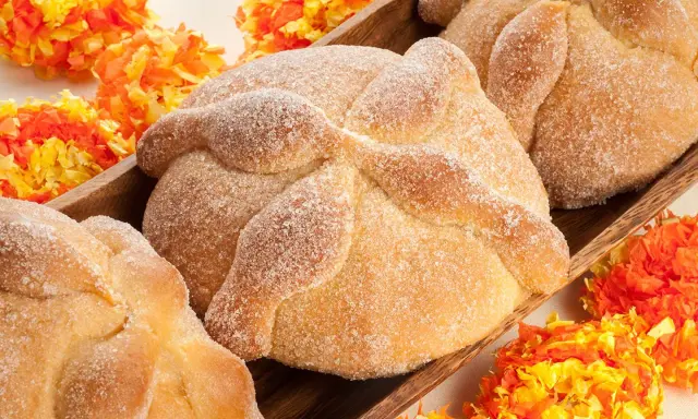 Pan de muerto, süßes Brot mit Zucker bestreut, liegt in einer Holzschale, umgeben von bunten Papierblumen.