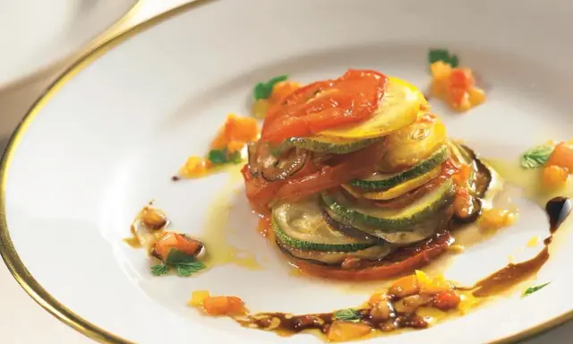 Ein kunstvoll geschichtetes Ratatouille-Rezept, inspiriert vom Film, mit Tomaten, Zucchini und Aubergine auf einem Teller mit goldenem Rand.