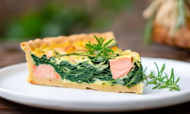 Ein Stück Lachs Spinat Quiche mit Rosmarin garniert auf einem weißen Teller.