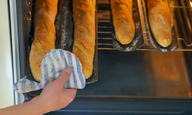 Hand mit Topflappen holt goldbraune Baguettes aus dem Ofen. Der Duft von frisch gebackenen Baguettes liegt in der Luft.