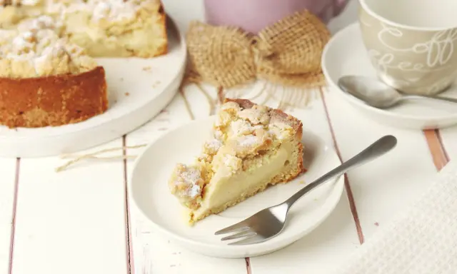 Ein Stück Streuselkuchen mit Pudding auf einem Teller, daneben eine Kaffeetasse und ein ganzer Kuchen.