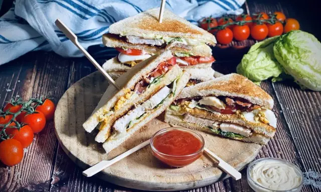 Ein gestapelter Club Sandwich mit Hühnchen, Speck, Ei und Tomaten, serviert mit Ketchup und Mayonnaise.