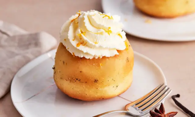 Ein köstlicher Baba au Rhum, gekrönt mit Schlagsahne und Orangenzesten, wartet auf einem Teller. Daneben liegen Sternanis und Vanilleschote.