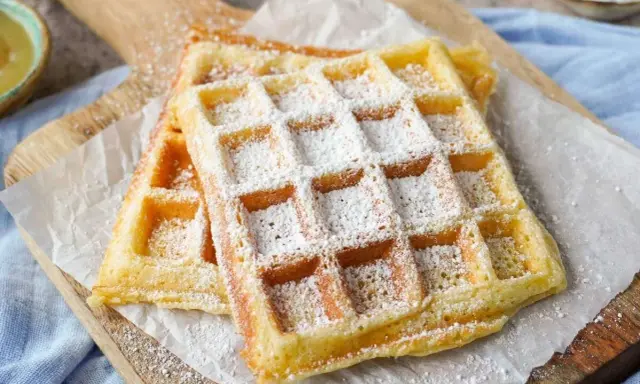 Zwei goldbraune belgische Waffeln, bestäubt mit Puderzucker, liegen auf Backpapier auf einem Holzbrett.