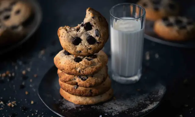 Ein Stapel amerikanischer Cookies mit Schokoladenstückchen, daneben ein Glas Milch. Ein Cookie ist angeknabbert.