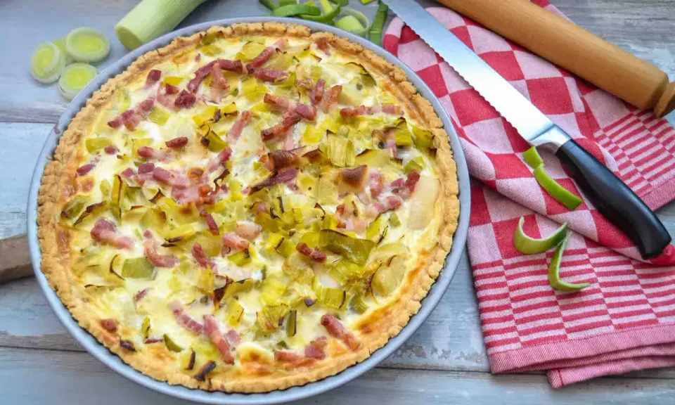 Herzhafte Quiche mit Lauch und Speck, perfekt für herzhafte Quiche Rezepte. Daneben liegen Lauch, ein Messer und ein Nudelholz.