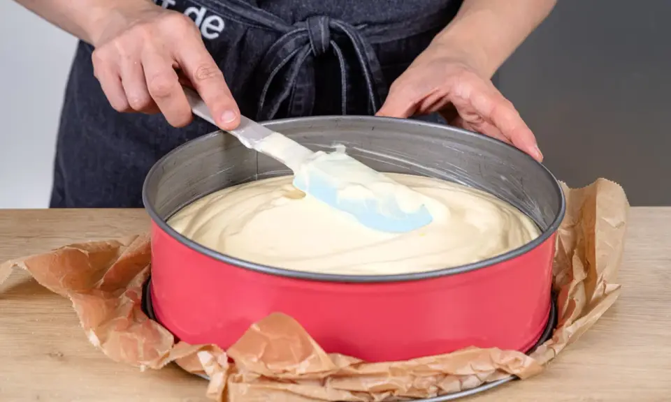 Hände verteilen den Teig für einen Joghurtkuchen mit 500 g Joghurt in einer roten Springform.
