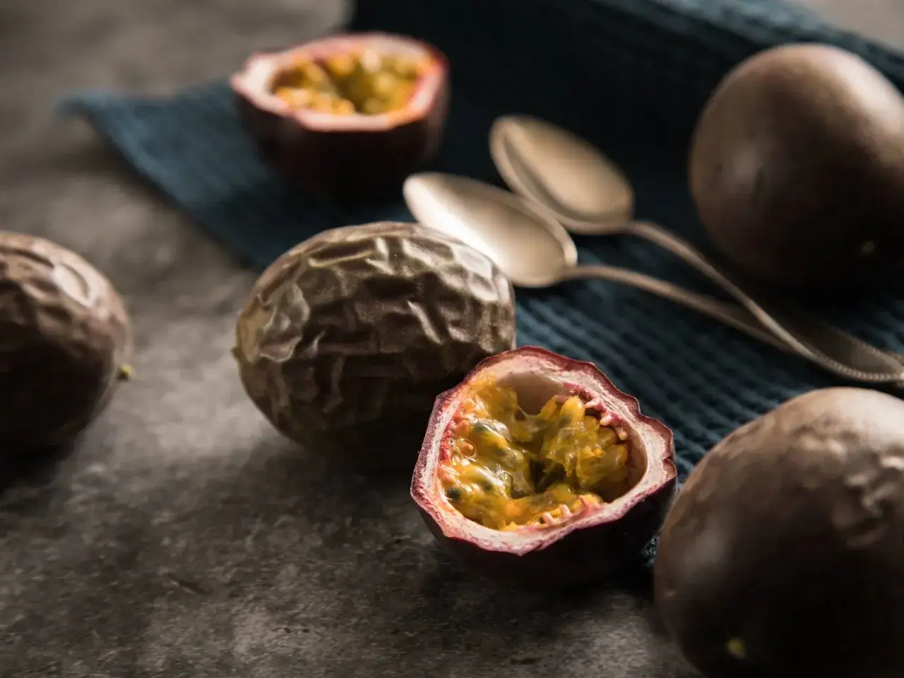 Reife Passionsfr&uuml;chte liegen auf einem dunklen Untergrund, eine ist aufgeschnitten und zeigt ihr saftiges Inneres. L&ouml;ffel liegen bereit, um das exotische Fruchtfleisch zu genie&szlig;en.