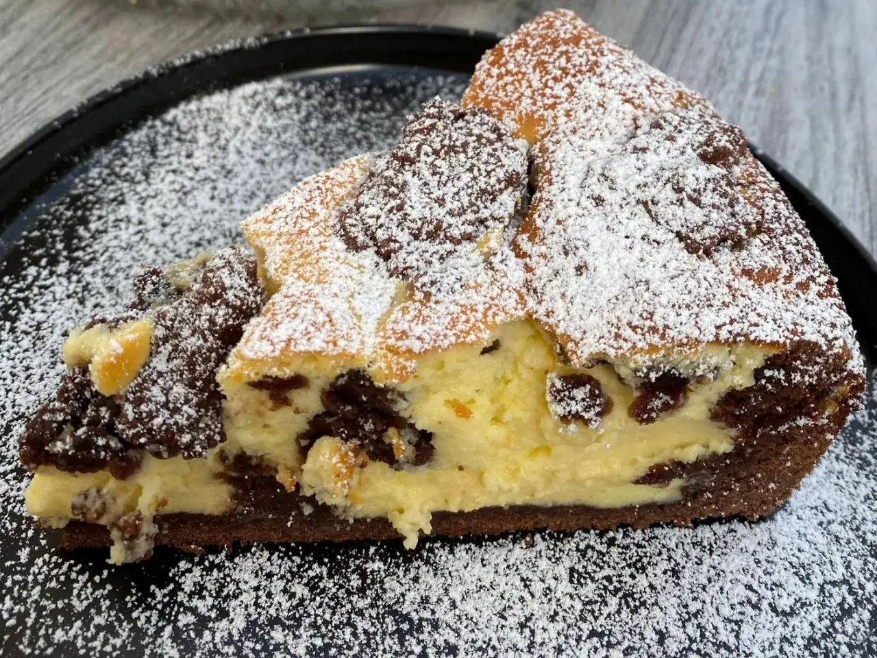 Ein saftiges St&uuml;ck versunkener Streuselkuchen mit Schokolade und Puderzucker best&auml;ubt, auf einem schwarzen Teller.