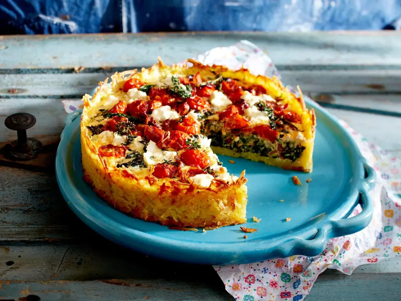 Herzhafte Quiche mit Spinat und Feta, belegt mit Kirschtomaten und Kr&auml;utern, auf einem blauen Teller.