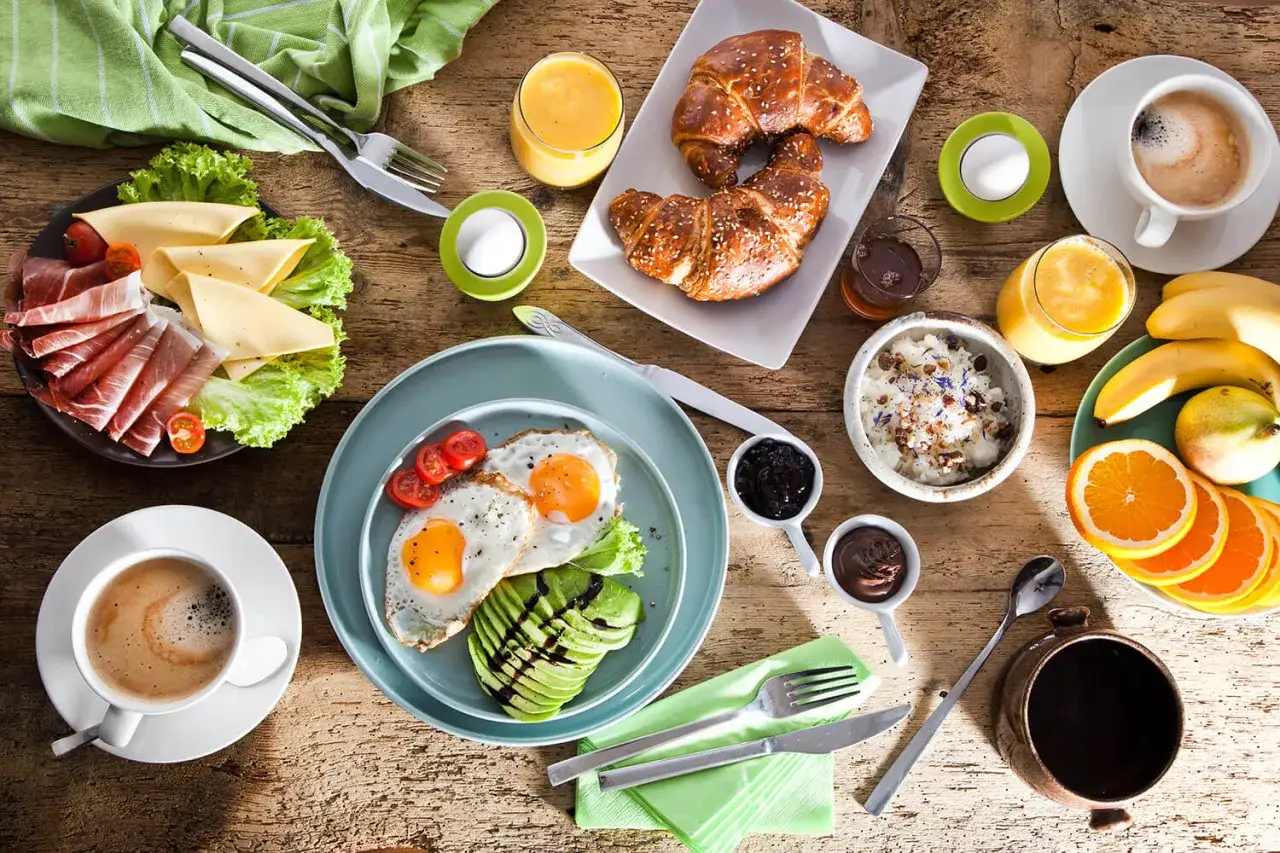 Leckere Brunch-Ideen: Spiegeleier mit Avocado, Schinken, K&auml;se, Croissants, M&uuml;sli, Obst und Kaffee auf Holztisch.