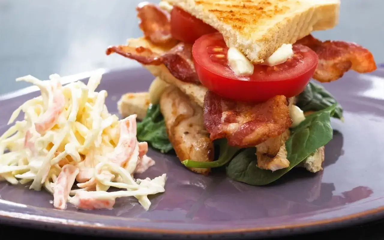 Ein saftiger Club Sandwich mit knusprigem Speck, Tomaten, H&auml;hnchen und Salat, serviert mit einem cremigen Krautsalat.