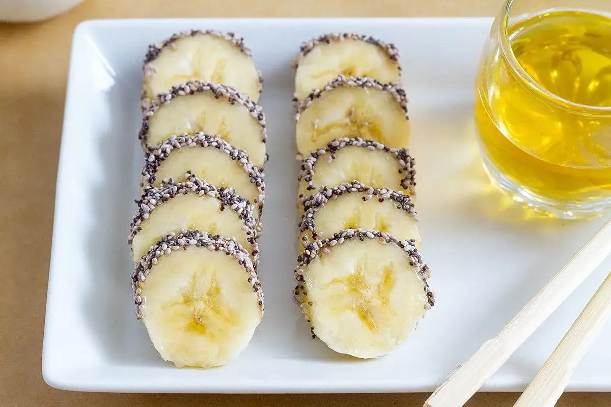 Bananenscheiben mit Chia-Samen und Honig, eine einfache Idee f&uuml;r gesunde Snack-Ideen. Daneben ein Glas mit gelbem Getr&auml;nk.