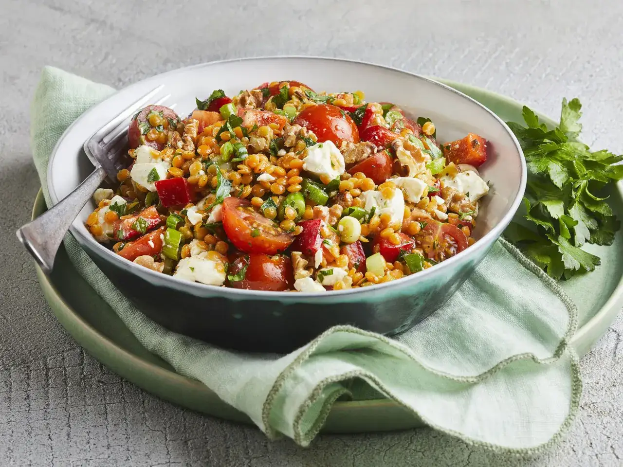 Ein bunter, gesunder rote Linsen Salat mit Tomaten, Feta und N&uuml;ssen, serviert in einer Sch&uuml;ssel auf einem gr&uuml;nen Teller mit Petersilie.