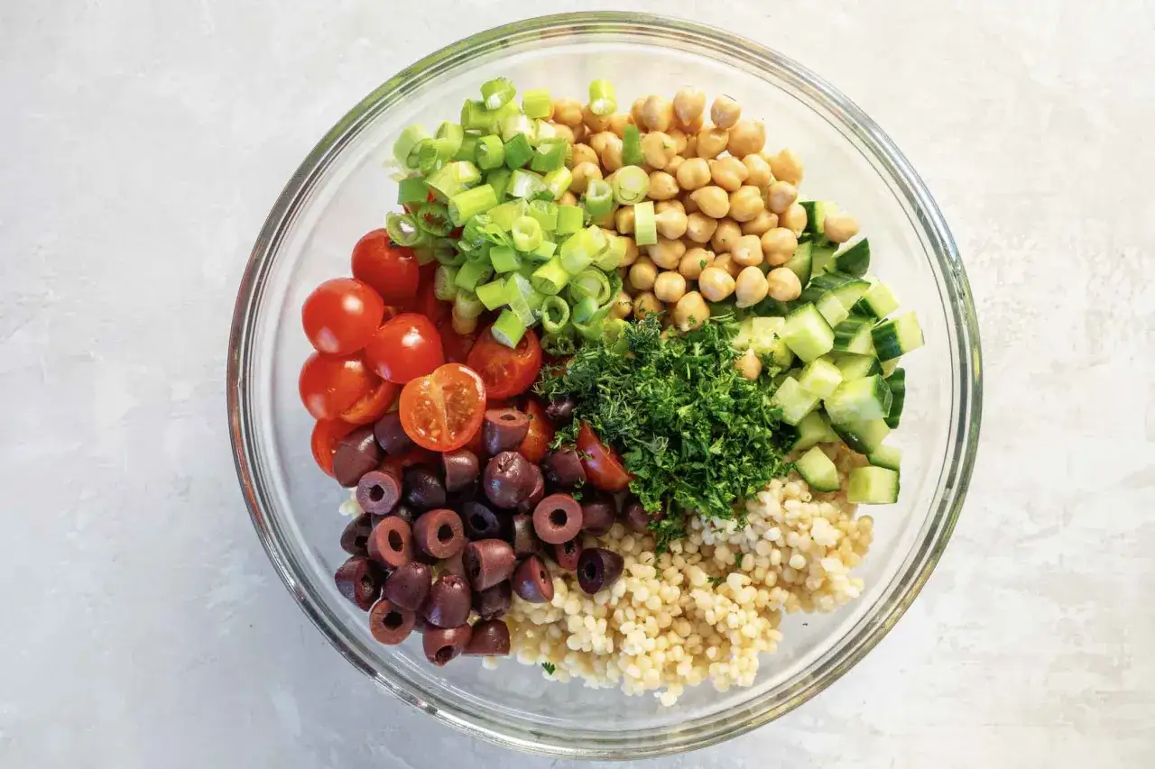 Bunte Zutaten für einen Couscous-Salat: Kichererbsen, Tomaten, Gurken, Oliven, Frühlingszwiebeln und Petersilie in einer Glasschüssel.