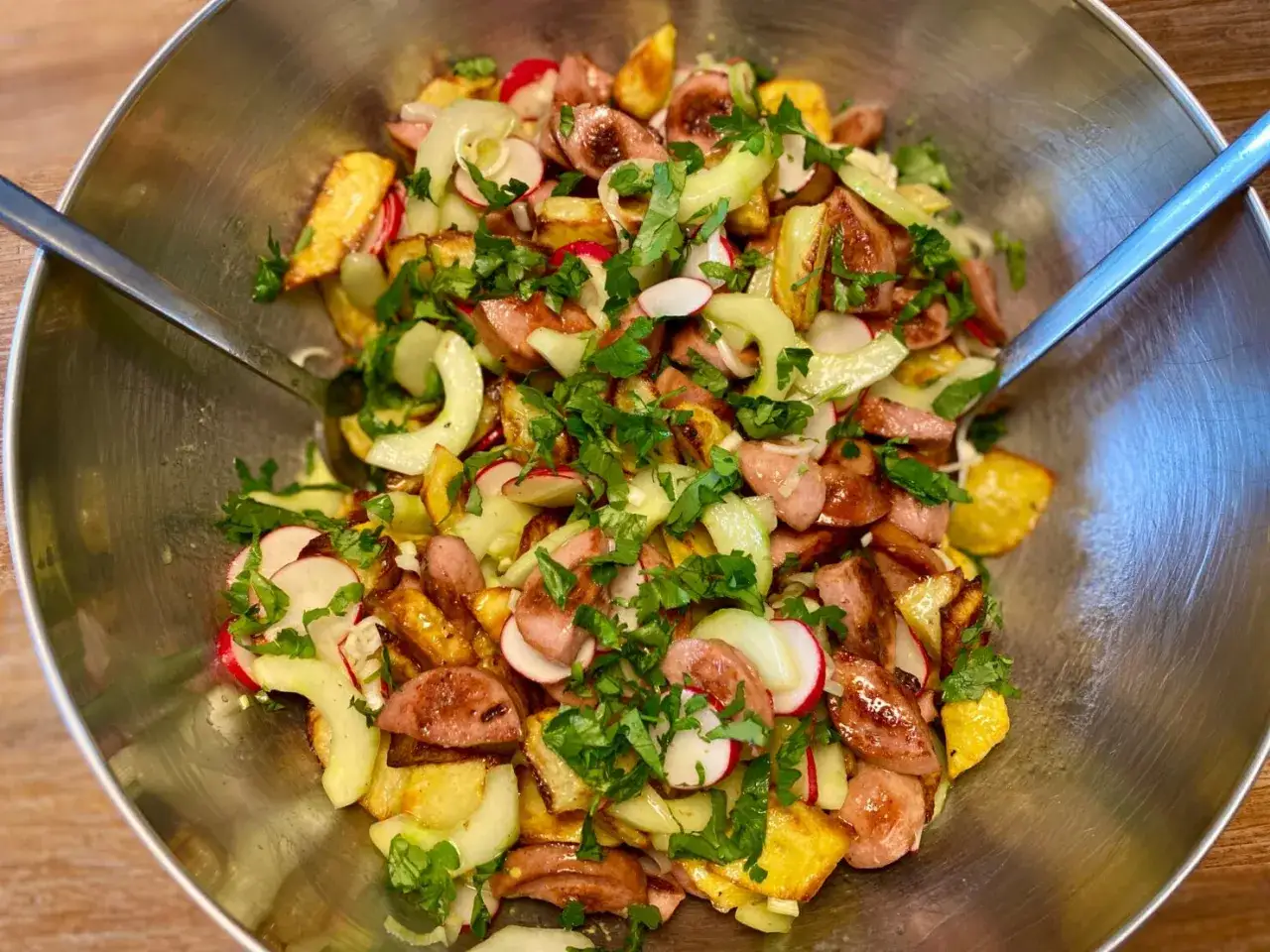 Ein herzhafter, crunchy Kartoffelsalat mit Wurstscheiben, Radieschen und Gurke, garniert mit frischer Petersilie in einer Edelstahlsch&uuml;ssel.