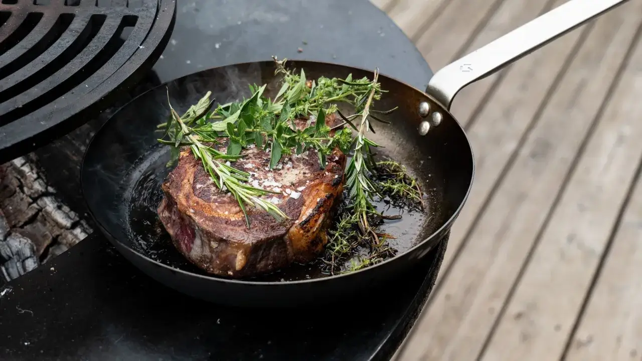 Ein saftiges Steak wird in einer Pfanne mit Rosmarin und Thymian richtig gebraten. Rauch steigt auf, w&auml;hrend das Fleisch brutzelt.
