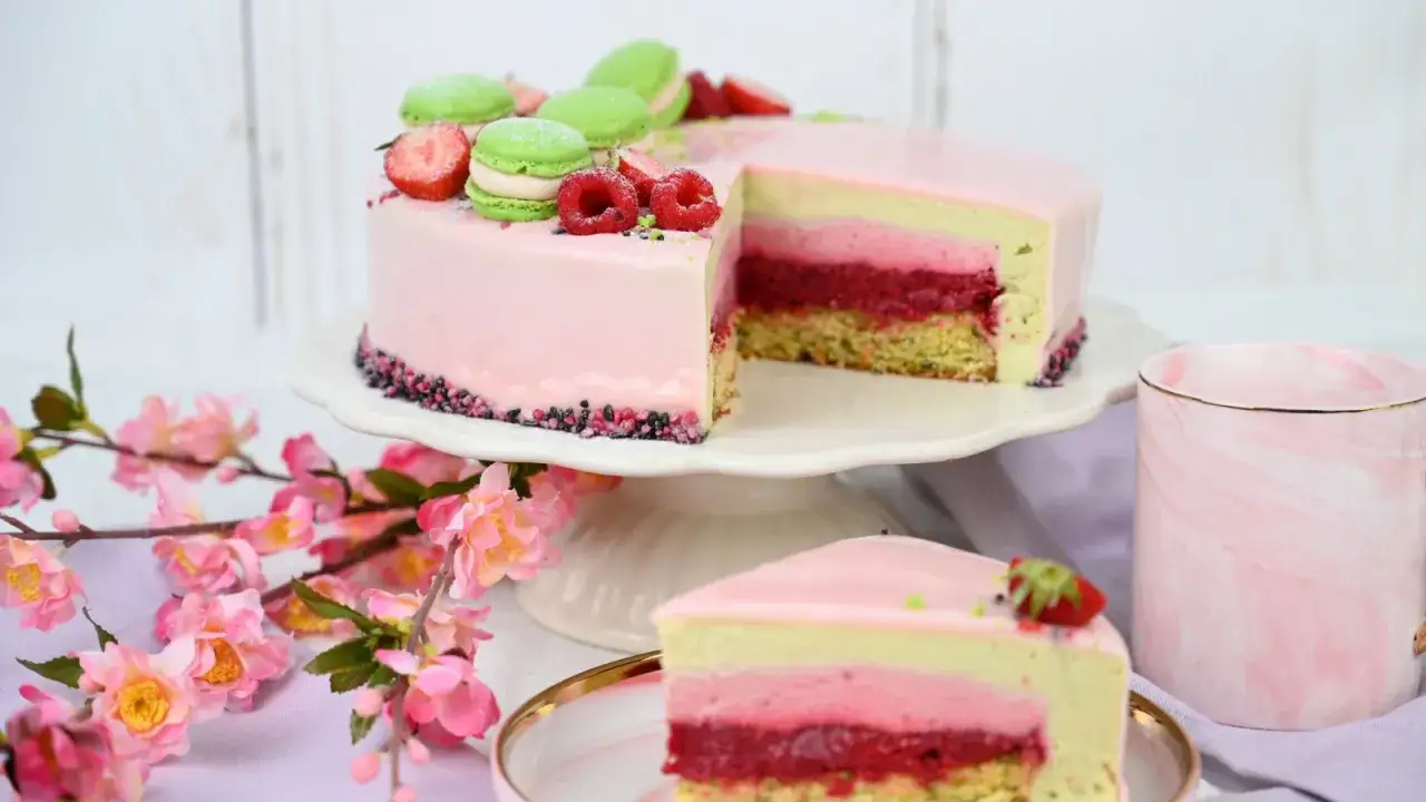 Eine Himbeer-Pistazien-Torte mit rosa Glasur, garniert mit Macarons und Beeren. Ein St&uuml;ck liegt auf einem Teller.