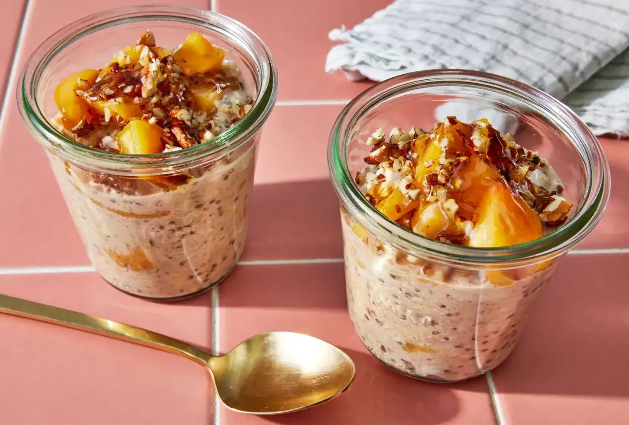 Zwei Gl&auml;ser mit Overnight Oats low carb, garniert mit Pfirsichst&uuml;cken, N&uuml;ssen und Sirup. Eine goldene L&ouml;ffel liegt daneben.