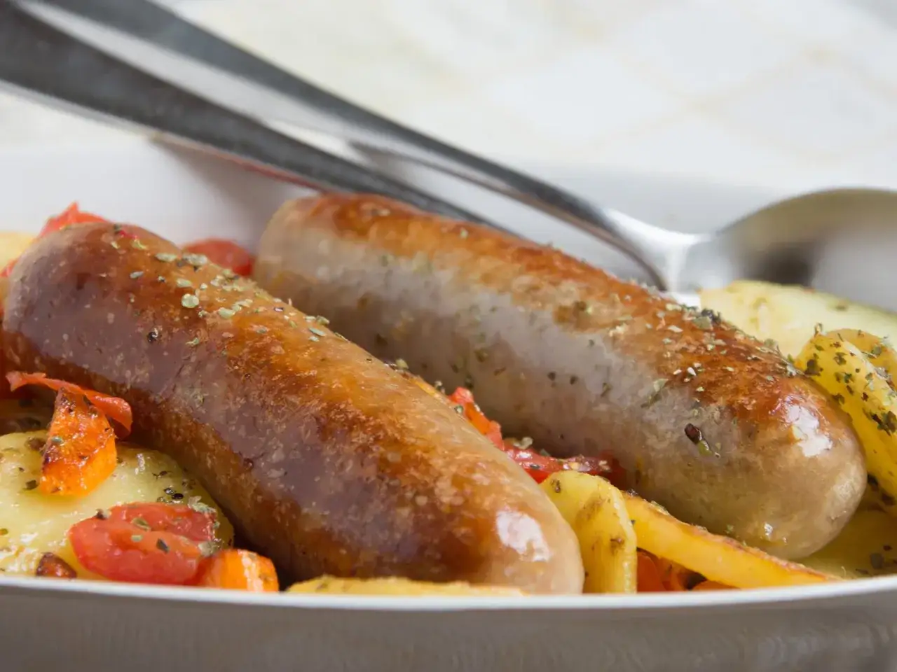 Zwei goldbraune Bratw&uuml;rste im Backofen mit Kartoffeln und Gem&uuml;se, appetitlich gew&uuml;rzt.