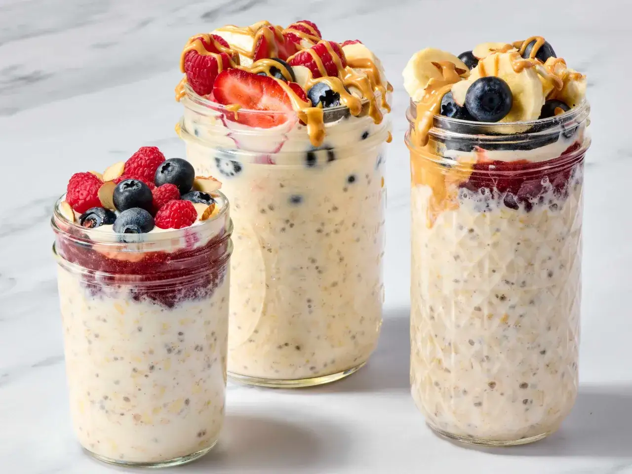 Drei Gl&auml;ser mit Overnight Oats, garniert mit Beeren, Bananen und Erdnussbutter. Perfekt f&uuml;r ein schnelles Fr&uuml;hst&uuml;ck.