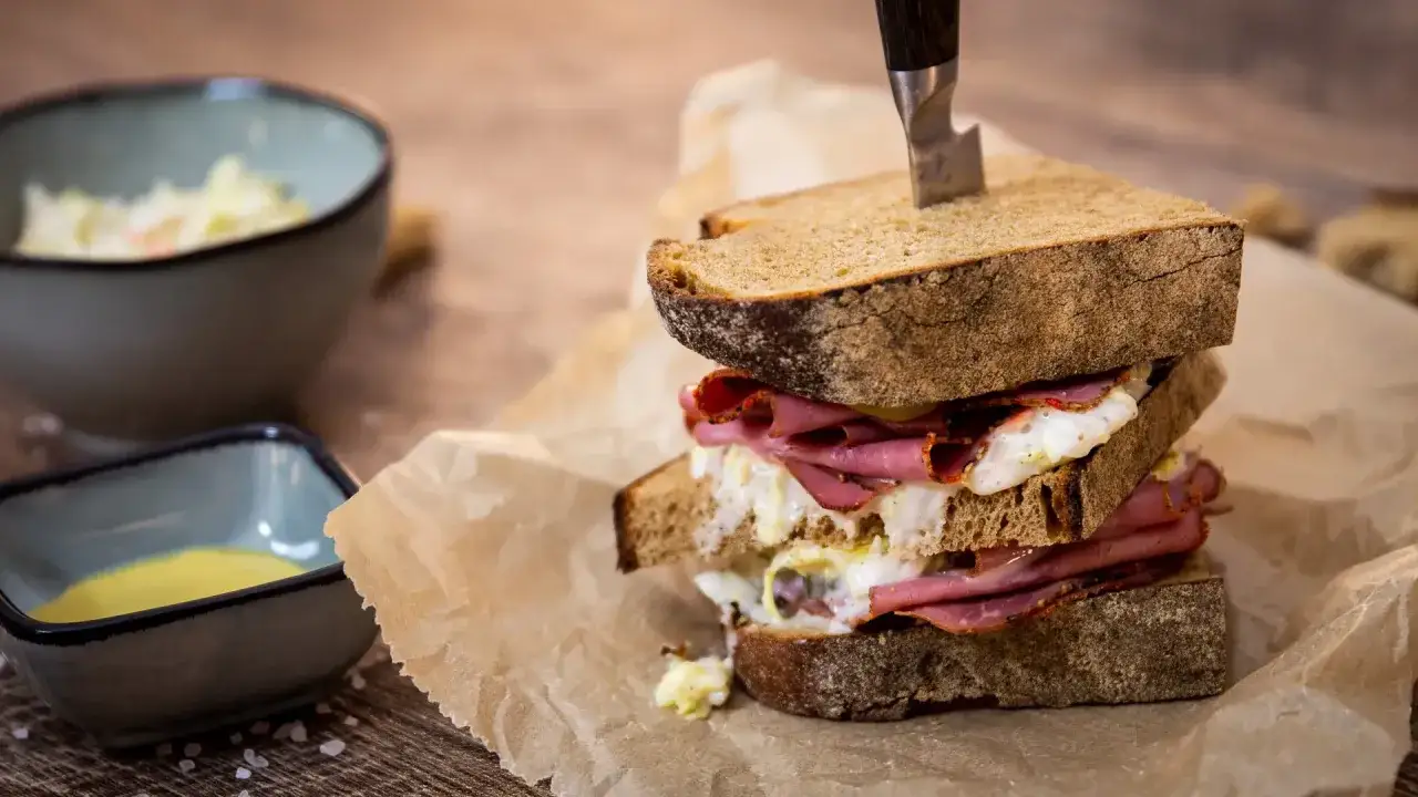 Ein &uuml;ppiges Pastrami-Sandwich mit Rindfleisch, Ei und Sauce, serviert auf rustikalem Brot. Daneben Sch&uuml;sseln mit Beilagen.