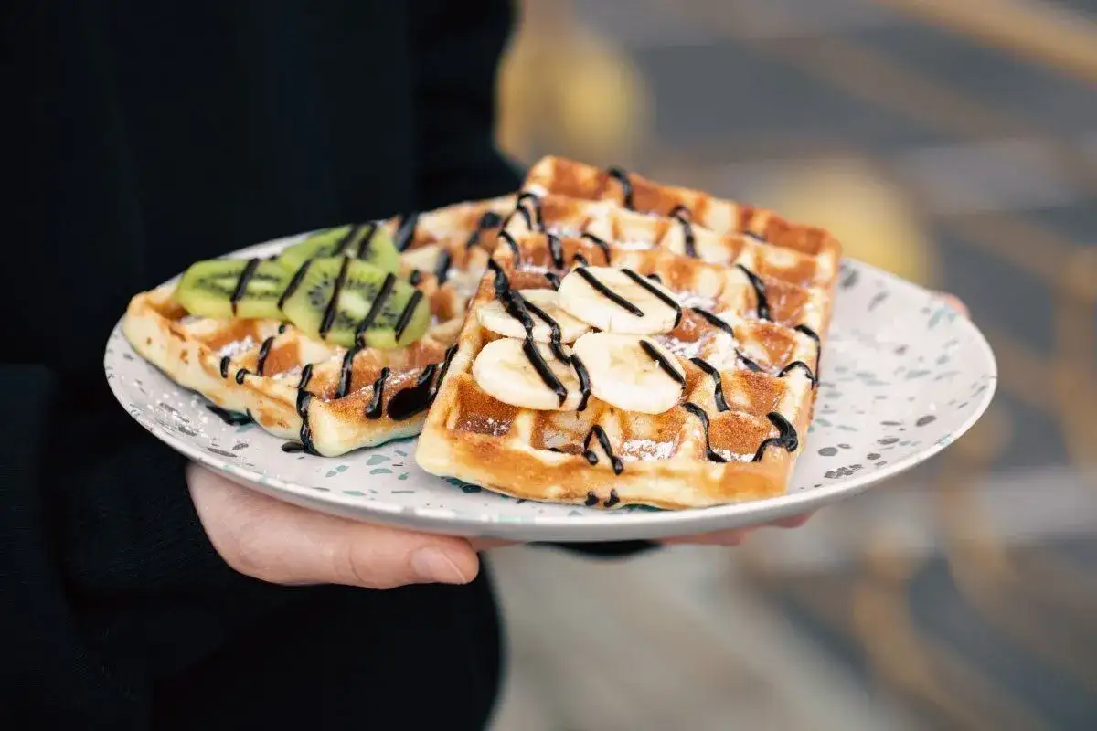 Zwei belgische Waffeln mit Bananenscheiben, Kiwist&uuml;cken und Schokoladensauce auf einem Teller.