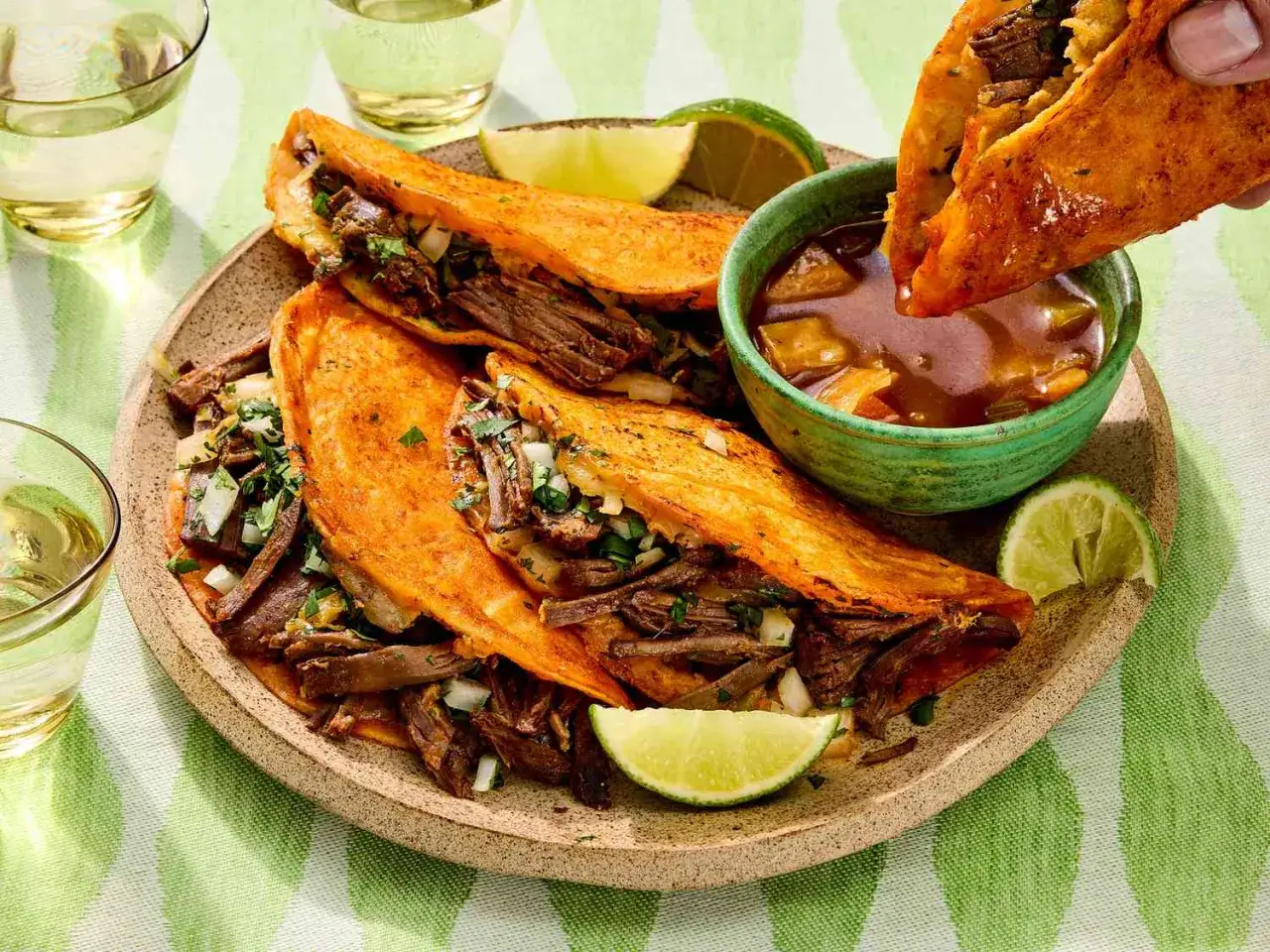 Saftige birria tacos mit Rindfleisch, Zwiebeln und Koriander, serviert mit einer w&uuml;rzigen Br&uuml;he und Limettenspalten. Ein Genuss!