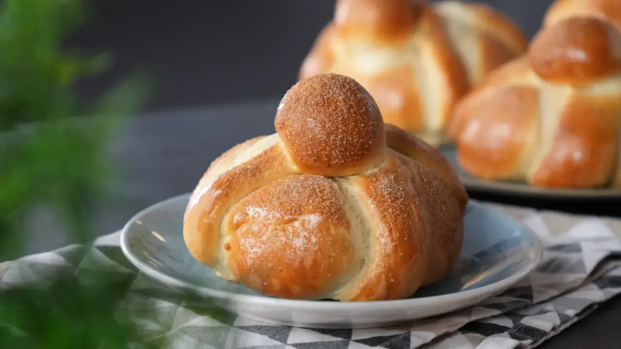 Ein goldbraunes Pan de Muerto, bestreut mit Zucker, auf einem blauen Teller. Im Hintergrund weitere s&uuml;&szlig;e Brote.