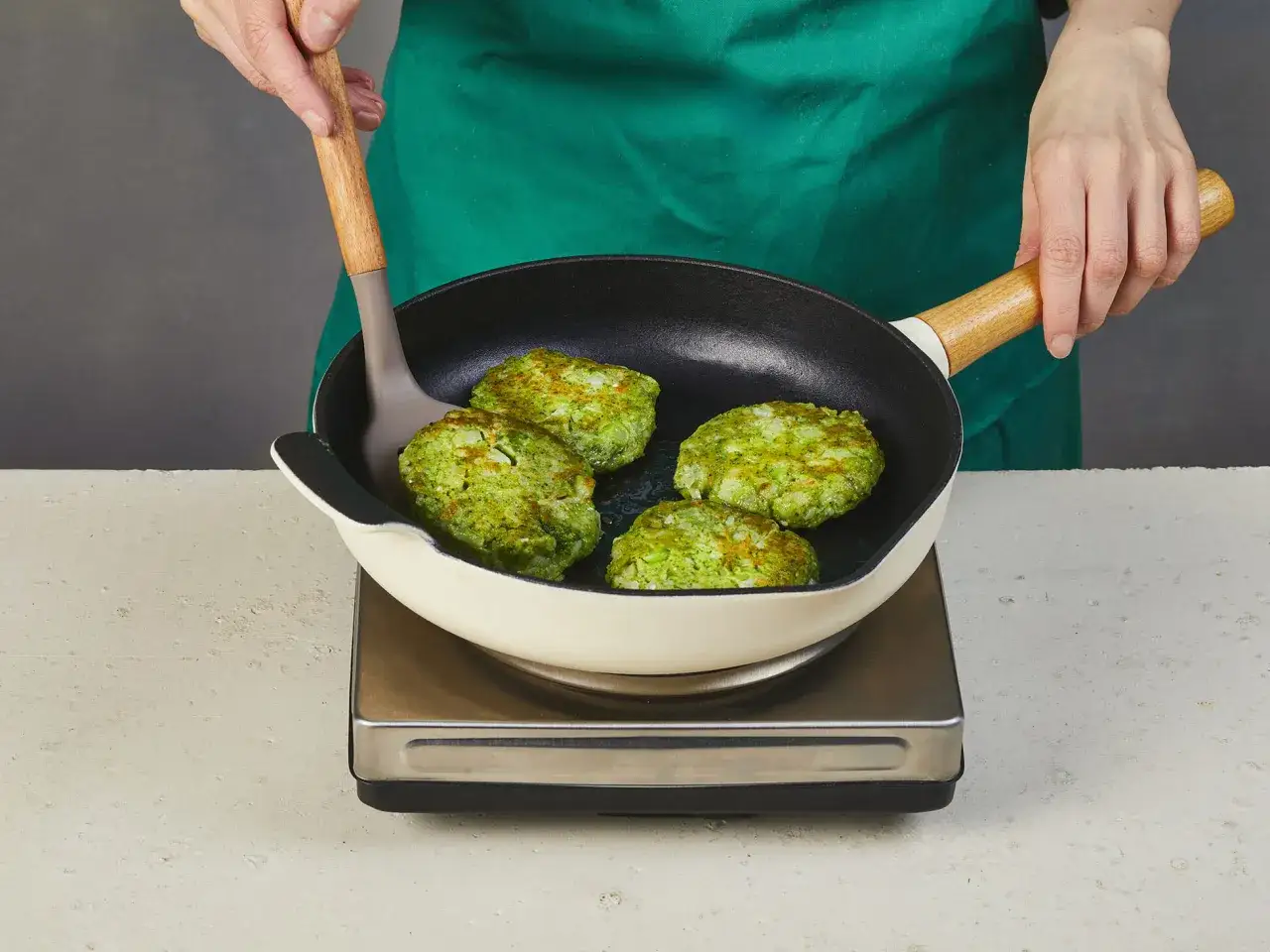 Gr&uuml;ne Brokkoli-Patties braten in einer Pfanne. Eine Hand mit einem Pfannenwender wendet die Patties.