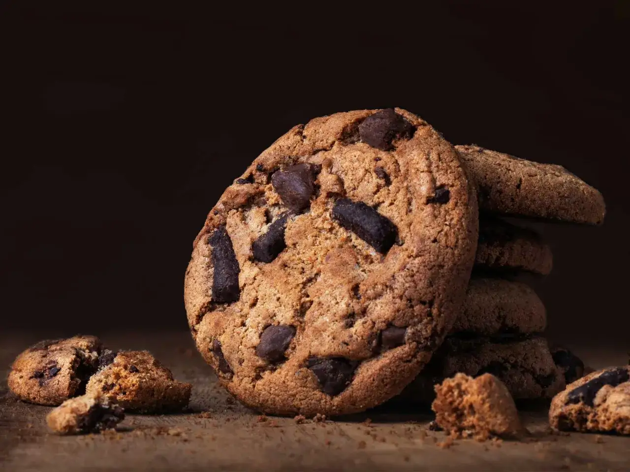 Ein Stapel saftiger american cookies mit Schokoladenst&uuml;ckchen auf einem Holztisch. Ein Cookie ist zerbrochen, Kr&uuml;mel liegen verstreut.