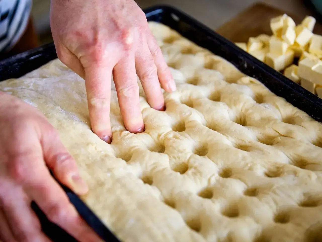 H&auml;nde dr&uuml;cken Vertiefungen in den Teig f&uuml;r ein Butterkuchen Rezept. W&uuml;rfel aus Butter liegen bereit.