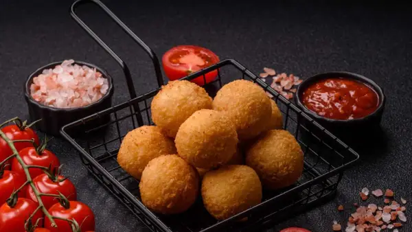 Goldbraune Arancini im Korb, bereit für ein köstliches arancini rezept. Dazu frische Tomaten, Salz und Dip.