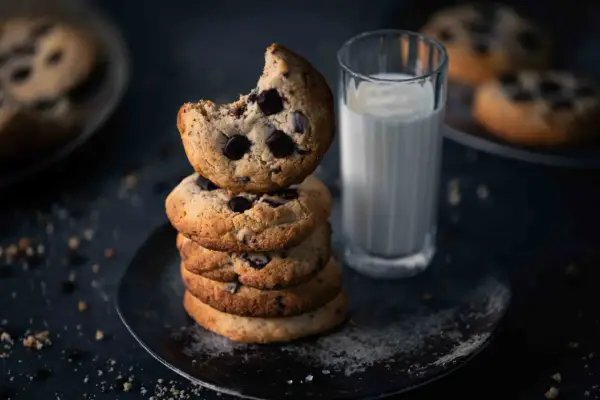 Ein Stapel amerikanischer Cookies mit Schokoladenstückchen, daneben ein Glas Milch. Ein Cookie ist angeknabbert.