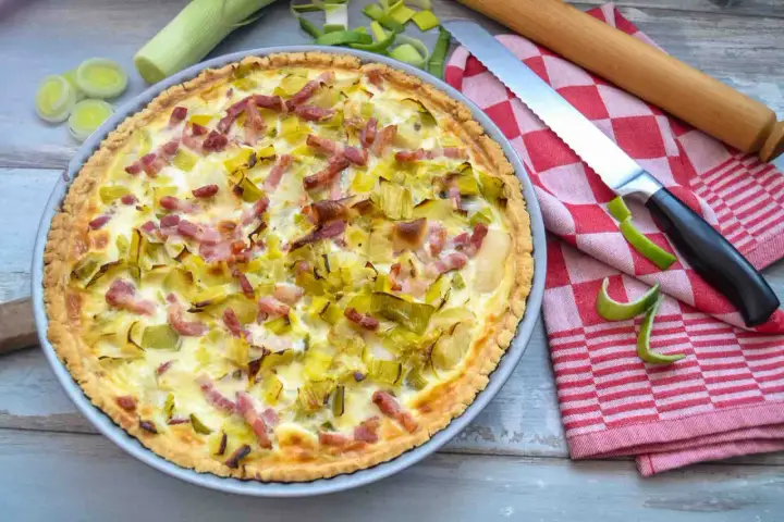 Herzhafte Quiche mit Lauch und Speck, perfekt für herzhafte Quiche Rezepte. Daneben liegen Lauch, ein Messer und ein Nudelholz.