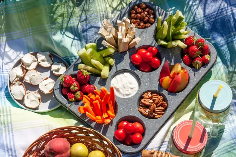 Leckere Picknick-Snacks: Erdbeeren, Gurken, Karotten, Nüsse, Tomaten, Pfirsiche und Brotscheiben mit Frischkäse auf einer Decke.